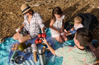 Çocuklar battaniyenin üzerinde piknik yapan bir aile. Güneşli bir günde rahat ve mutlu bir ortamda meyve ve atıştırmalık paylaşıyorlar..