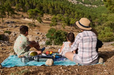 Üç kişilik aile dağlarda sakin bir piknik yapıyor. Meyve ve atıştırmalıklarla bir battaniyeye oturup doğal manzaranın tadını çıkarıyorlar. Aile zamanı, doğa ve rahatlama kavramları için ideal..