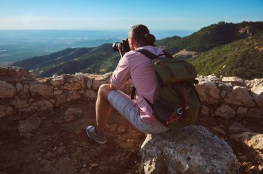 Sırt çantalı bir fotoğrafçı, gün batımında kayalık bir tepede oturan güzel bir dağ manzarasının fotoğraflarını çekiyor..