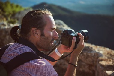 Kendini adamış bir fotoğrafçı elinde kamerayla güzel bir dağ manzarasının fotoğraflarını çekiyor. Sahne yaratıcılığı ve doğa fotoğrafçılığını çağrıştırıyor..
