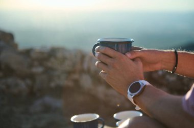 Metal bir fincan kahveyi tutan ellerin yakın çekimi, güneşli sakin bir sabahın tadını çıkarıyor. Rahatlama, açık hava macerası ve sükunet temaları için mükemmel..