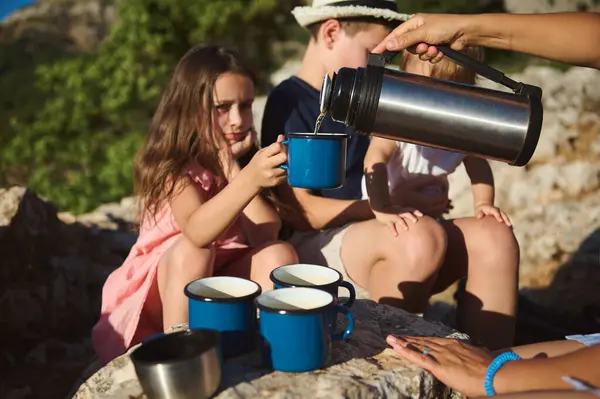 Stock image A family enjoys a camping trip in the mountains, with children being served hot beverages in blue mugs from a thermos, amid a natural, scenic environment.