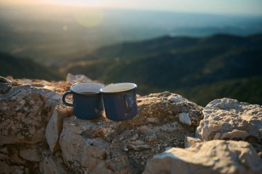 İki mavi diş mineli kahve fincanı kayalık bir dağın tepesinde dinleniyor ve nefes kesici bir gün doğumu manzarası var. Mükemmel bir huzur, macera ve doğal güzellik anı..