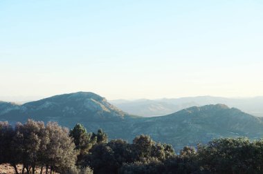 Önünde bereketli ağaçlar ve berrak mavi gökyüzünün altındaki uzak tepelerin olduğu huzurlu bir dağ manzarası..