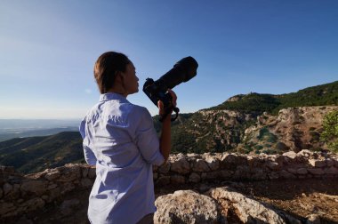 Profesyonel bir kamera tutan bir kadın dağların ve vadilerin manzarasını yakalar. Açık bir günde fotoğraf, seyahat ve doğa keşfi kavramı.