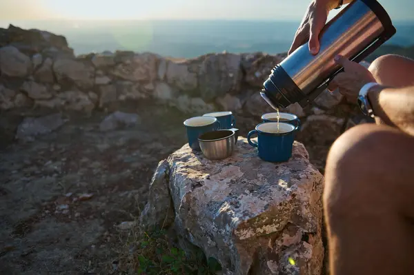 stock image Person pouring coffee from a thermos into metal cups on a rocky surface, set against a scenic background, capturing the essence of outdoor adventure and relaxation.