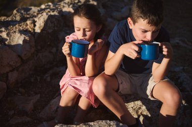 İki çocuk kayalık arazide oturup sıcak güneş ışığı altında mavi bardaklardan gelen sıcak içeceklerin tadını çıkararak bir anlık boş zamanı yakalıyorlar..