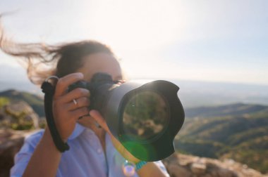 Güneşli, dağlık bir arazide profesyonel bir kamerayla fotoğraf çeken bir fotoğrafçının yakın çekimi. Rüzgâr saçlarında esiyor, doğayı yakalama anını vurguluyor..