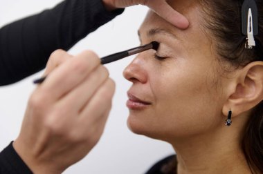 Makyaj sanatçısının bir kadının göz kapağına göz farı sürerken yakın plan fotoğrafı. Kadın, güzellik sürecinde rahat ve hoşnut görünüyor..