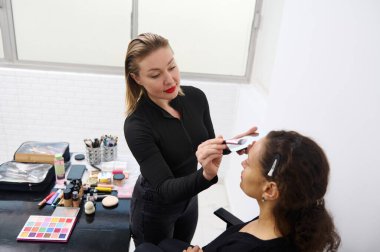 A professional makeup artist applies eyeshadow to a client's eyelids. Various makeup products and tools are seen on the table in a bright and clean studio. clipart