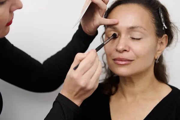 stock image Close-up of a professional makeup artist applying eye shadow to a woman's eyelids, creating a smooth and flawless look. The woman appears calm and relaxed during the beauty session.