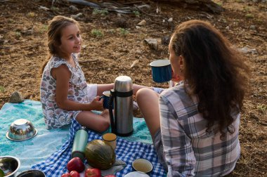 Anne ve kızı dışarıda piknik yaparken sıcak bir şeyler içiyorlar. Sahne, doğal ortamdaki bir bağ ve rahatlama anını yakalar..