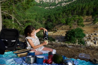 Genç kız manzaralı bir ormanda yaz pikniğinin tadını çıkarıyor. Piknik malzemeleri ve doğa ile çevrili bir termostan su dolduruyor..