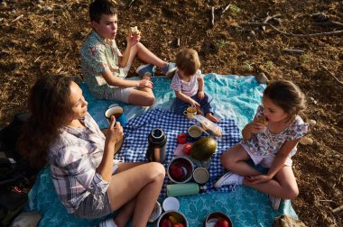 Çocuklarla dışarıda piknik yapan ve abur cubur yiyen bir aile. Sahne, doğadaki neşeli bir anı yakalıyor, aile bağlarını ve açık hava aktivitelerini vurguluyor..