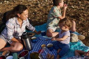 Neşeli bir aile güneşli bir günde dışarıda piknik yapıyor, yiyecek ve içecekleri paylaşıyor. Sahne mutluluk ve birliktelik dolu..