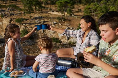Neşeli bir aile açık havada piknik yapıyor. İki çocuk ve anneleri fincanla kadeh kaldırırken üçüncü çocuk atıştırmanın tadını çıkarıyor..