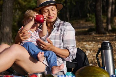Anne ve çocuk ormanda birlikte vakit geçiriyorlar taze meyvelerle pikniğin tadını çıkarıyorlar. Çocuk kırmızı bir elma tutuyor, sağlıklı yaşam tarzını ve aile bağını simgeliyor..