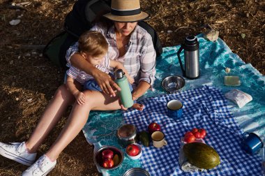 Bir anne ve çocuğu dışarıda bir battaniyede piknik yapıyorlar. Yiyecek ve doğa ile çevrelenmiş olarak birlikte huzurlu ve güneşli bir günün tadını çıkarıyorlar..