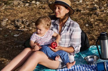 Anne ve bebek doğada piknik battaniyesinde oturup güneşli bir günün tadını çıkarıyorlar. Çocuk kucağında otururken anne bir elma tutar. Etrafları piknik malzemeleriyle çevrili..
