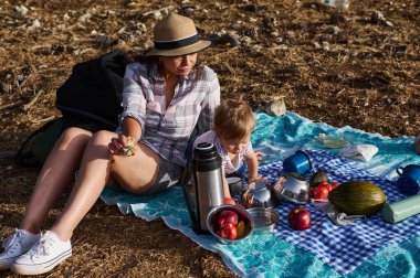 Bir anne ve çocuk dışarıda piknik yapıyor. Çeşitli meyveler ve termosla bir battaniyenin üzerine otururlar. Sahne sıradan, huzurlu ve doğal güneş ışığıyla dolu..