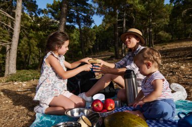 Bir anne ve iki çocuğu ormanda rahatlatıcı bir piknik yapıyorlar. Aile güneşli bir günde yemeğini paylaşıyor ve doğanın tadını çıkarıyor..