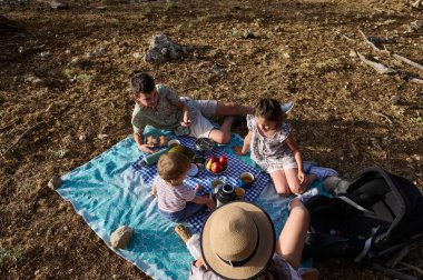 Bir grup çocuk dışarıda battaniyenin üzerinde piknik yapıyorlar. Güneşli bir günde içecek ve meyve veriyorlar, doğanın ve açık havanın tadını çıkarıyorlar..