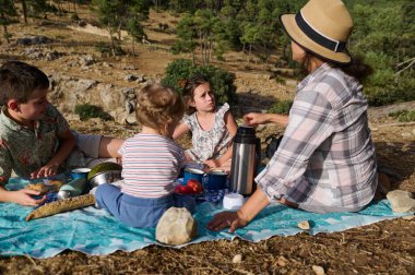 Bir anne ve çocukları doğa manzaralı bir ortamda piknik yapıyorlar. Çocuklar etraflarında çeşitli piknik malzemeleriyle bir battaniyenin üzerinde oturuyorlar..