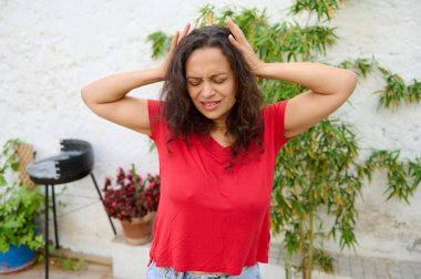 Kırmızı gömlekli bir kadın dışarıda dikilirken hayal kırıklığı ve rahatsızlık içinde başını tutuyor. Arka plan yeşil bitkilerle dolu bir ev bahçesi. Stres ve endişe hissi veriyor..