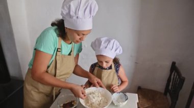 Bir anne ve kızı evde birlikte yemek pişiriyorlar. İkisi de şef şapkası ve önlüğü takıyor. Mutfakta kaliteli vakit geçiriyorlar..