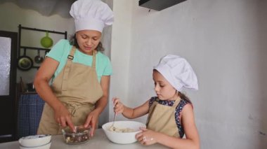 Bir anne ve kızı mutfakta yemek yaparken birbirlerine bağlanıyorlar, ikisi de aşçı şapkası ve önlüğü takıyorlar. Anne malzemeleri hazırlarken kız malzemeleri bir kasede karıştırıyor..