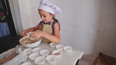 Şef şapkası ve önlüğü takan genç bir kız evde kek hamuru hazırlıyor. Pişirmedeki yaratıcılığını ve neşesini gösteriyor. Mutfak sanatı devam ediyor.