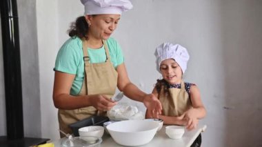 Bir anne ve kız mutfakta birlikte yemek pişirmekten zevk alır, uyumlu önlükler ve şef şapkaları giyerek. Malzemeleri hazırlayıp pişirme sürecine bağlarlar..