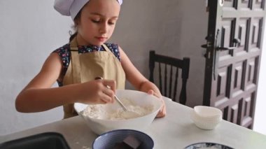Önlük ve şef şapkası takan genç bir kız evde yemek pişirirken malzemeleri kaseye karıştırıyor. Mutfaktaki işine odaklanmış..