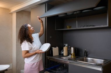 Günlük kıyafetli bir kadın mutfak dolabını düzenliyor, elinde beyaz bir tabak tutuyor ve modern bir mutfak ortamında bulaşıkları düzenliyor..