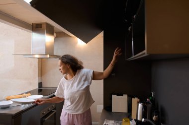 Modern bir mutfak ortamında günlük giysiler giymiş bir kadın kahvaltı hazırlıyor. Tezgahın üstüne tabak ve ekmek yerleştirir, rahat ve rahat bir atmosfer ekler..