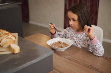 Genç bir kız ahşap bir masada oturmuş kahvaltıda bir kase mısır gevreği yiyor. Yakınlardaki tezgahta dilimlenmiş ekmek görülebiliyor. Sıcak bir kahvaltı ortamı yaratıyor..