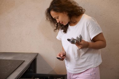 Beyaz tişörtlü bir kadın modern bir mutfakta gümüş çatal bıçak takımı düzenlerken görülüyor. İşine odaklanmış görünüyor..