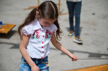 Kırmızı işlemeli beyaz gömlek ve mavi kot pantolon giyen genç bir kız güneşli bir günde bir ip oyuncağıyla dışarıda oynuyor. Odaklandı ve oyun zamanının tadını çıkarıyor..