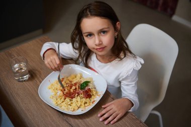 Tahta bir masada oturan genç bir kız yanında bir bardak suyla domates sosu ve peynirli makarnanın tadını çıkarıyor. Mutlu ve odaklanmış görünüyor..