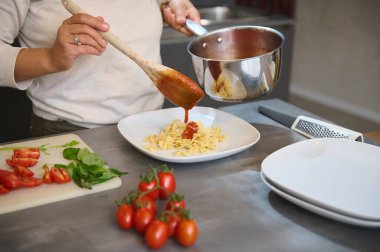 Modern bir mutfakta taze domates ve fesleğenle ev yapımı makarnanın üzerine taze domates sosu döken birinin yakın çekimi..