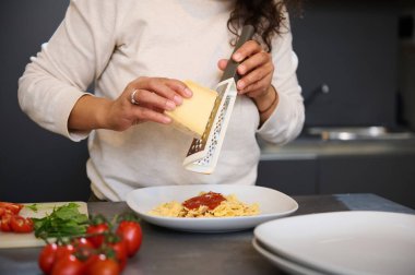 Close-up of a woman grating cheese onto a plate of pasta in a contemporary kitchen. Fresh tomatoes, spinach, and other ingredients are visible on the counter. clipart