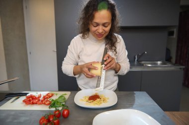 A woman grating cheese over a plate of pasta in a modern kitchen with fresh ingredients like tomatoes and basil on the counter, focusing on cooking and meal preparation. clipart