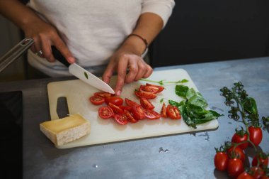Bir parça peynirle taze kiraz domatesleri ve fesleğen yapraklarını dilimleyen birinin yakın plan çekimi, sağlıklı beslenme ve yemek hazırlığını vurguluyor.