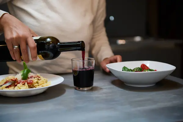 stock image A close-up of a hand pouring red wine into a glass, accompanied by a pasta and salad meal, creating a cozy home dining atmosphere.