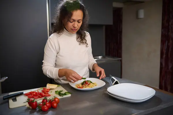 stock image Woman in a modern kitchen garnishing a pasta dish with basil. Fresh ingredients like tomatoes and cheese are on the countertop.