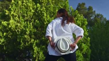 Beyaz gömlekli ve şapkalı bir kadın yemyeşil bir parkta yürüyor yemyeşil ağaçlarla dolu, güneşin ve doğanın tadını çıkarıyor..