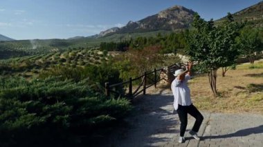 Zeytin ağaçları, dağlar ve doğa ile çevrili güzel bir kırsal alanda neşeyle dans eden bir adam berrak mavi gökyüzünün altında huzurlu manzaranın tadını çıkarıyor..