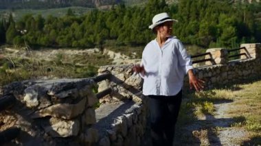 Beyaz gömlekli ve şapkalı bir kadın kırsal kesimdeki yemyeşil tepelerin ve taş duvarların önünde neşeyle dans ediyor. Sahne özgürlük ve neşeyi temsil eder..