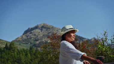 Şapka takan sakin bir kadın, arka planda dağ manzaralı doğayı kucaklıyor. Güneşli bir günde doğal bir ortamda huzur ve rahatlığı yakalamak..