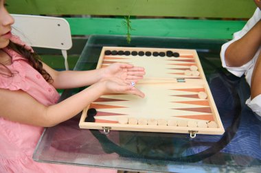Açık hava ortamında yeşil arka planlı cam bir masada tavla oynayan iki çocuk. Odaklanma tahtada ve oyuncuların ellerinde..